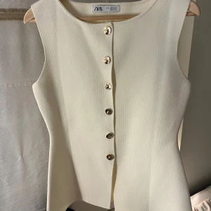 Vit väst från Zara - Snygg vit väst från Zara med eleganta guldknappar framtill. Västen har en stilren design och är ärmlös, vilket gör den perfekt för lager-på-lager. Passar bra till både vardag och festliga tillfällen.