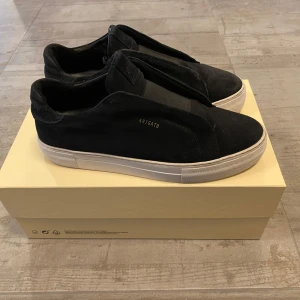 Svarta sneakers från Axel Arigato - Snygga svarta sneakers från Axel Arigato med en stilren design. Skorna har en vit sula och är tillverkade i mocka. Perfekta för en minimalistisk look. Passar till det mesta och är smidiga att ta av och på eftersom att dem inte har skosnören man behöver knyta. Man får såklart med allt på bilderna. Dem är normala i storleken.