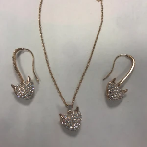 Strass Guldhalsband och örhängen med hjärtan - Elegant set med halsband och örhängen i guld. Både halsbandet och örhängena har hängande hjärtan dekorerade med glittriga stenar. Perfekt för att ge en touch av glamour till din outfit.  Helt ny 