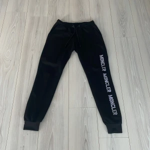 Svarta mjukisbyxor från Moncler - Snygga svarta mjukisbyxor från Moncler med logga längs benet och en liten patch på baksidan. Byxorna har snörning i midjan och muddar vid bensluten för en bekväm passform.