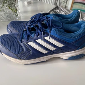 Blå Adidas sneakers - Snygga blå Adidas sneakers med vita ränder och snörning. Skorna har en sportig design med en bekväm passform och är perfekta för träning och vardagsbruk . De har en slitstark sula och andningsbart material för extra komfort.