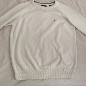 Gant tröja  - Säljer en stilren vit stickad tröja från Gant med diskret logga på bröstet. Tröjan har långa ärmar och ribbade muddar vid ärmslut och nederkant. Perfekt för en klassisk och avslappnad look.