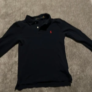 Svart pikétröja långarmad från Ralph Lauren !! PRISET EJ HUGGET I STEN!! - Snygg svart pikétröja från Ralph Lauren med klassisk krage och knappar. Storlek 10/12 passar även xxs säljer för att den är för liten och inte kommit till användning på länge OBS!! PRISET INTE HUGGET I STEN