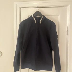 Mörkblå jacka från Stone Island - Säljer en snygg mörkblå jacka från Stone Island med dragkedja och luva. Jackan har ett stilrent märke på ärmen och är perfekt för en cool och avslappnad stil.