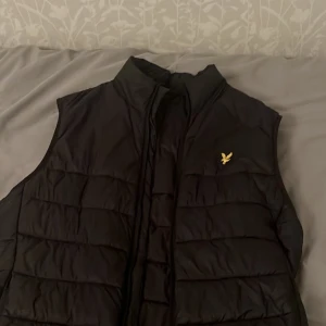 Svart dunväst med dragkedja - Snygg svart dunväst med dragkedja och en liten guldfärgad logga på bröstet. Perfekt för kyligare dagar och ger en stilren look. Västen har en hög krage för extra värme.