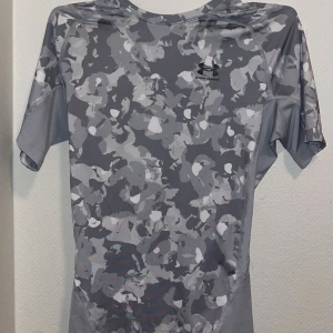 Grå camouflagemönstrad t-shirt från Under Armour - Snygg grå camouflagemönstrad t-shirt från Under Armour. Perfekt för träning med korta ärmar och en bekväm passform. Tillverkad i ett lätt och andningsbart material.
