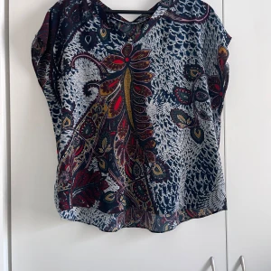 Mönstrad blus med blommor - Säljer en färgglad blus med ett unikt blommönster i rött, blått och gult. Blusen har korta ärmar och en lös passform, perfekt för en bohemisk stil. Materialet är lätt och luftigt, vilket gör den bekväm att bära.