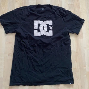 Svart t-shirt från DC - Säljer en svart t-shirt från DC med logga i vitt på bröstet. T-shirten har en klassisk passform och korta ärmar, perfekt för en avslappnad stil. Passar 170-185cm
