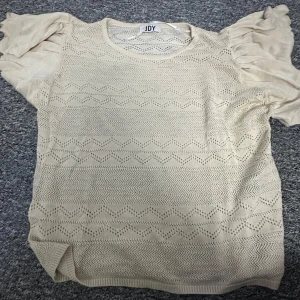Beige mönstrad topp från JDY - Säljer en söt beige stickad topp från JDY med korta volangärmar. Toppen har ett fint hålmönster som ger en luftig känsla. Perfekt för en stilren och avslappnad look. Passar bra till både jeans och kjol. 🌟