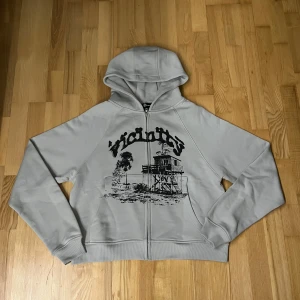 Grå hoodie från Vicinity - Säljer en grå hoodie från Vicinity med dragkedja och tryck av en stuga och träd på framsidan. Perfekt för en avslappnad stil. Hoodien har långa ärmar och en bekväm passform.