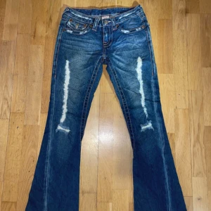 Blå bootcut jeans från True Religion - Snygga blå bootcut jeans från True Religion med slitna detaljer på framsidan. Klassisk femficksdesign och knappar med märkets logga. Perfekta för en avslappnad stil. W25