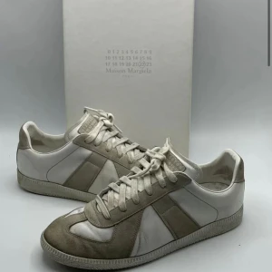 Sneakers från Maison Margiela - Stilrena sneakers från Maison Margiela i en kombination av vitt och beige. Skorna har en klassisk design med snörning och är tillverkade i en mix av skinn och mocka. Perfekta för en avslappnad men ändå elegant look.
