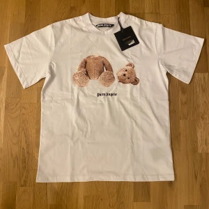 Vit t-shirt med nalletryck från Palm Angels - Säljer en vit t-shirt från Palm Angels med ett unikt nalletryck på framsidan. T-shirten har korta ärmar och Palm Angels-loggan i svart på baksidan. Perfekt för en stilren och trendig look.