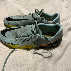 Nike fotbollsskor i ljusblått och gult - Säljer ett par Nike fotbollsskor i ljusblått med svarta detaljer och en gul sula. Skorna har snörning och är designade för optimal prestanda på planen. Perfekta för fotbollsspelare som vill ha stil och funktion.