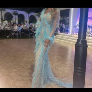 Vlora Kaltrina Klänning från kosovo - Säljer en fantastisk långklänning i ljusblått med fjäderdetaljer och glittriga inslag. Klänningen har en v-ringad design och långa ärmar i ett transparent material. Perfekt för en glamorös kväll! Nypris 9000kr. Använt bara en gång