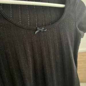 Svart ribbad topp från Bik Bok - Säljer en snygg svart ribbad topp från Bik Bok. Den har korta ärmar och en söt liten rosett vid halsringningen. Endast använd 1 gång 