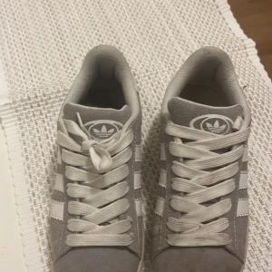 Grå Adidas sneakers - Säljer ett par grå Adidas sneakers i mocka med vita detaljer. De har den klassiska tre-randiga designen på sidorna och Adidas-loggan på plösen och hälen. Skorna har snörning och ser ut att vara i bra skick, perfekta för vardagsbruk.