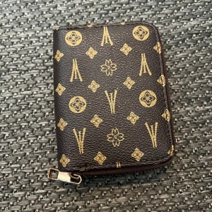 Svart plånbok med guldmönster - Säljer en snygg brun plånbok med ett coolt guldmönster. Den har en dragkedja runtom och flera fack inuti för kort och kontanter. Perfekt för att hålla ordning på dina grejer!