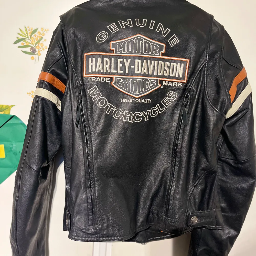 Säljer en svart skinnjacka från Harley-Davidson med coola detaljer. Jackan har långa ärmar och dragkedja framtill. Den har också snygga orange och vita ränder på ärmarna och ett stort Harley-Davidson-märke på ryggen. Köpte i New York. Passa storlek s/m. Jackor.