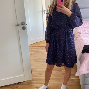 Blommig klänning från Esprit - Säljer en superfin blommig klänning från Esprit. Den är blå med små röda och rosa blommor och har en knytning i midjan för en snygg passform. Klänningen har långa ärmar och en v-ringad hals. Perfekt för både vardag och fest!