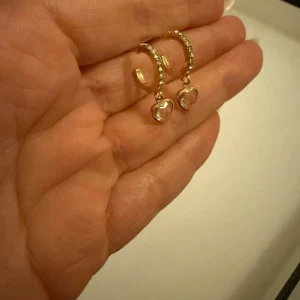 Guldörhängen med hjärtformade hängen - Eleganta guldörhängen med gnistrande stenar och hjärtformade hängen. Perfekta för att ge en romantisk touch till din stil. Örhängena har en klassisk design med en modern twist.