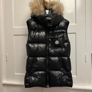 Svart dunväst från Moncler - Snygg svart dunväst från Moncler med en fluffig pälsdetalj på huvan. Huvan går att ta av se bild. Västen har en dragkedja framtill och en praktisk bröstficka med knapp. Perfekt för kyligare dagar när du vill hålla stilen. Storlek Small. Använd en gång så gott som ny!