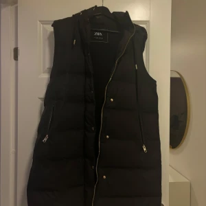 Svart dunväst från Zara - Snygg svart dunväst från Zara med dragkedja och huva. Perfekt för kyliga dagar och har praktiska fickor med dragkedjor. Västen är stilren och lätt att matcha med olika outfits.