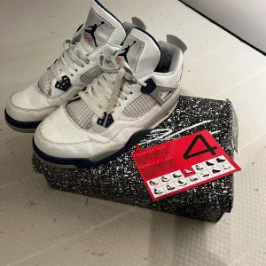 Air Jordan 4 Retro i vit och mörkblå - Säljer ett par klassiska Air Jordan 4 Retro i vit och mörkblå färg. Skorna har en stilren design med snörning och detaljer i grått. Perfekta för dig som älskar sneakers. Det har tyvärr en trasig sak på sidan därför säljer jag billigt 