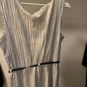 Silverfärgad ärmlös klänning - Snygg silverfärgad klänning med plisserat tyg och ärmlös design. Klänningen har en v-ringad hals och ett svart bälte i midjan som ger en fin silhuett. Perfekt för en glittrig look!