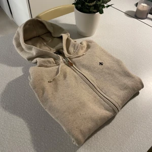 Beige hoodie från Morris - Säljer en snygg beige hoodie från Morris med dragkedja och huva. Tröjan har ribbade muddar och en liten broderad detalj på bröstet. Shysst att använda på vintern. Perfekt för en avslappnad stil. Säljer då jag inte använder den så mycket. 1700kr nypris!! Hör av dig vid intresse!
