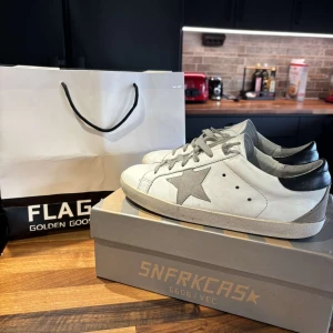 Golden Goose sneakers med stjärndetalj - Snygga vita sneakers från Golden Goose med en grå stjärna på sidan och svarta detaljer vid hälen. Skorna har snörning och en klassisk design som passar perfekt för en trendig look. Passar en storlek större stora i storlek. Passar 41 topp skick utan några defekter.
