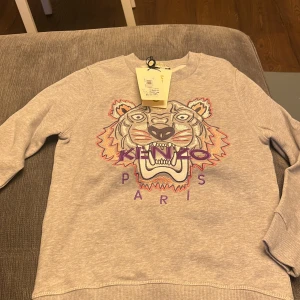 Helt oanvänd med prislapp grå sweatshirt från Kenzo med tigerbroderi - Säljer en grå sweatshirt från Kenzo med ett stort, färgglatt tigerbroderi på framsidan. Tröjan har långa ärmar och rund halsringning. Perfekt för en cool och avslappnad stil.
