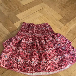 Röd mönstrad kjol - Charmig röd kjol med ett detaljerat mönster i vitt och svart. Kjolen har en elastisk midja för bekväm passform och har inne shorts . Perfekt för en färgglad look!