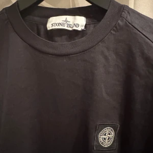 Svart långärmad t-shirt från Stone Island - Snygg svart långärmad t-shirt från Stone Island med deras ikoniska logga på bröstet. Perfekt för en stilren och avslappnad look. Tillverkad i ett bekvämt material som passar bra till vardags.
