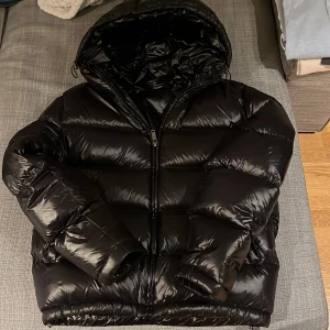 Svart pufferjacka med huva från Zara  - Säljer en svart pufferjacka med huva från Zara. Jackan har en glansig yta och dragkedja framtill. Perfekt för kyliga dagar när du vill hålla dig varm och stilren.