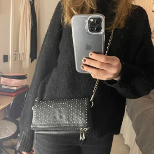 Svart axelväska med nitar - Snygg svart axelväska med silverfärgade nitar och kedja från even&odd. Perfekt för att ge din outfit en edgy touch. Väskan har en dragkedja för säker förvaring av dina saker. Den viks på mitten och stängs med två knappar. Tyvärr så har den lite defekter då kedjan/axelbandet och dragkedjan är en aning rostiga, men kedjan/axelbandet kan lätt bytas ut.🩵för med bilder eller frågor, skriv privat.