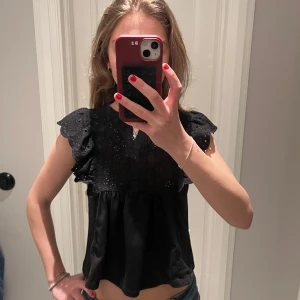 Svart blus med volangdetaljer - Säljer en svart blus med volangdetaljer på axlarna från MANGO. Blusen har en lös passform och är perfekt för både vardag och fest, passar perfekt nu till sommarn! Den är ärmlös och har en rund halsringning. Storleken är xs men skulle säga att den är mer som en S och passar även M