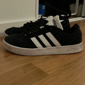Svarta sneakers från Adidas - Snygga svarta sneakers från Adidas med de klassiska tre vita ränderna på sidan. Skorna har en vit sula och snörning framtill. Perfekta för en avslappnad stil. Tvättas innan den skickas 