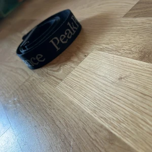 Svart bälte från Peak Performance - Säljer ett svart bälte från Peak Performance med vit text. Bältet har en robust svart spänne med logotyp. Perfekt för att ge en sportig touch till din outfit. Har även små repor här och var men ska det ska inte förstöra dess prestanda!