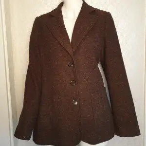 Snygg brun kavaj från Lindex med klassisk design och tre knappar framtill. Tillverkad i ett melerat tyg med diskreta färginslag. Perfekt för en stilren look.