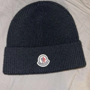Svart stickad mössa från Moncler - Snygg svart stickad mössa från Moncler med ett broderat märke framtill. Perfekt för att hålla värmen under kyliga dagar.