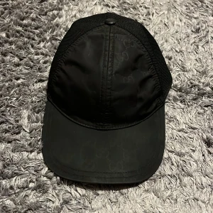 Svart keps från Gucci - Snygg svart keps från Gucci med justerbar passform. Kepsen har ett diskret mönster och nätpaneler baktill för ventilation. Perfekt för en stilren look.