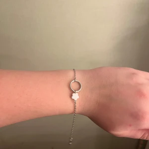 Silverfärgat armband med blomma - Elegant silverfärgat armband med en liten vit blomma som hänge. Armbandet har en tunn kedja och en justerbar längd med karbinhake för enkel användning. Perfekt för att lägga till en subtil touch av stil till din outfit.