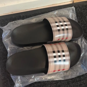 Burburry tofflor - Snygga svarta sandaler med klassiskt rutigt mönster i beige, svart och rött. Perfekta för en avslappnad stil. 