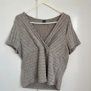 Grå ribbad omlottopp från SHEIN - Säljer en stilren grå ribbad omlottopp från SHEIN. Toppen har korta ärmar och en v-ringad design som ger en elegant look. Perfekt för både vardag och speciella tillfällen.