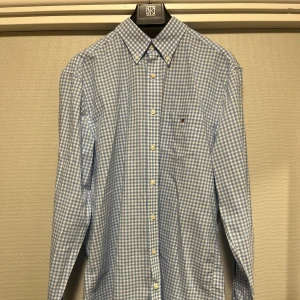 Blå och vit rutig skjorta från GANT - Säljer en klassisk blå och vit rutig skjorta från GANT i storlek L. Skjortan har en button-down krage och en bröstficka med GANT-logga. Perfekt för en stilren look!