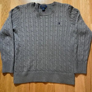 Grå kabelstickad tröja från Ralph Lauren - Säljer en stilren grå kabelstickad tröja från Ralph Lauren med det klassiska broderade logotypen på bröstet. Tröjan har en rund halsringning och långa ärmar, perfekt för kyligare dagar. Passar både till jeans och chinos för en avslappnad look.