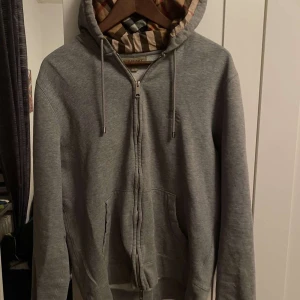 Grå hoodie från Burberry - Säljer en stilren grå hoodie från Burberry med klassiskt rutigt mönster i huvan. Tröjan har dragkedja framtill och justerbara snören vid huvan. Perfekt för en avslappnad och trendig look. Äkta !!