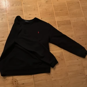 Svart tröja från Polo Ralph Lauren - Säljer en stilren svart tröja från Ralph Lauren med det klassiska logotypbroderiet i rött på bröstet. Tröjan har långa ärmar och en bekväm passform. Perfekt för en avslappnad och snygg look. 