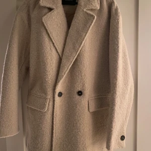 En kortare Beige kappa från Studio - Säljer en stilren beige kappa från Studio i storlek S. Kappan har en klassisk design med dubbelknäppning och långa ärmar. Perfekt för kyligare dagar med sin mysiga och varma känsla. 🧥✨